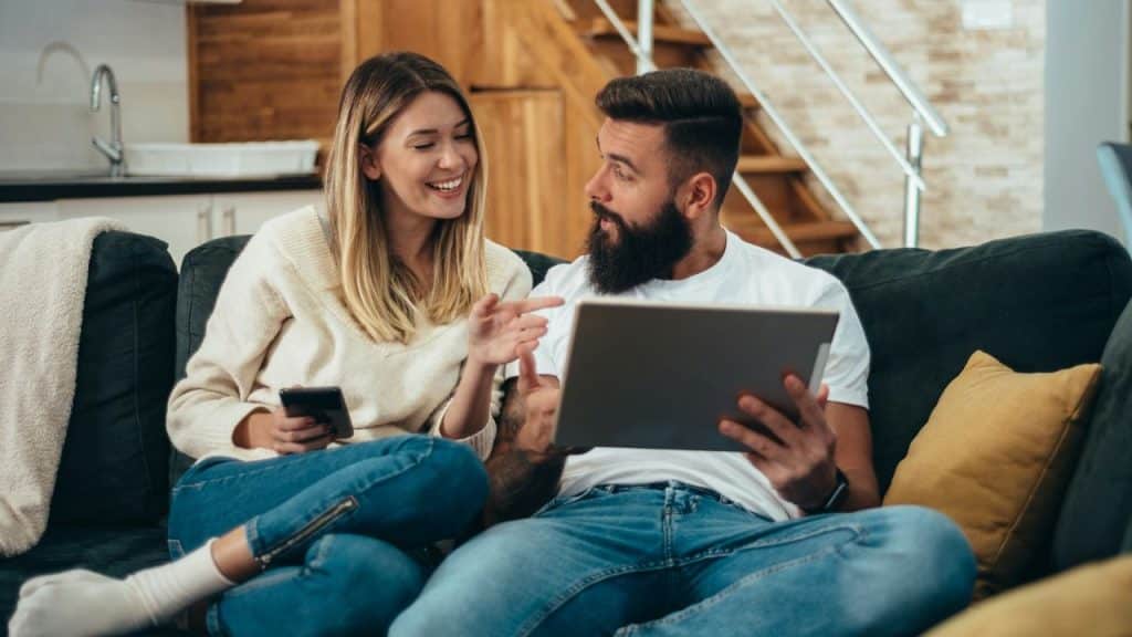 A smiling woman looks at a man holding a tablet while sitting on a couch.