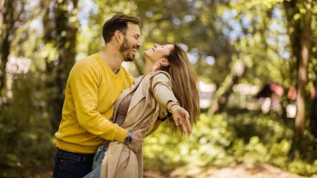 A smiling man holds a laughing woman, playfully dipping her in a park.
