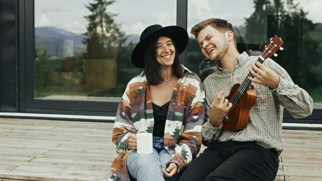 A joyful couple sits outside; the man plays a ukulele while the woman laughs.