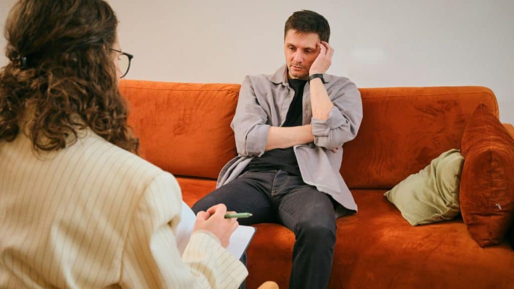 A thoughtful man sits on an orange couch, listening to a woman taking notes.