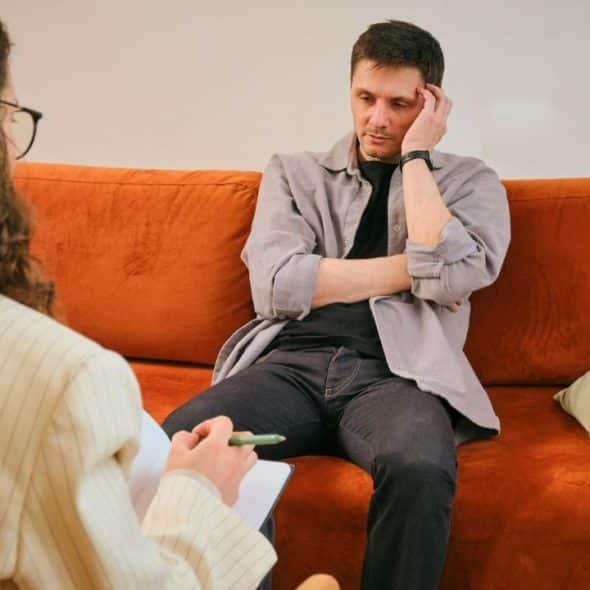 A thoughtful man sits on an orange couch, listening to a woman taking notes.