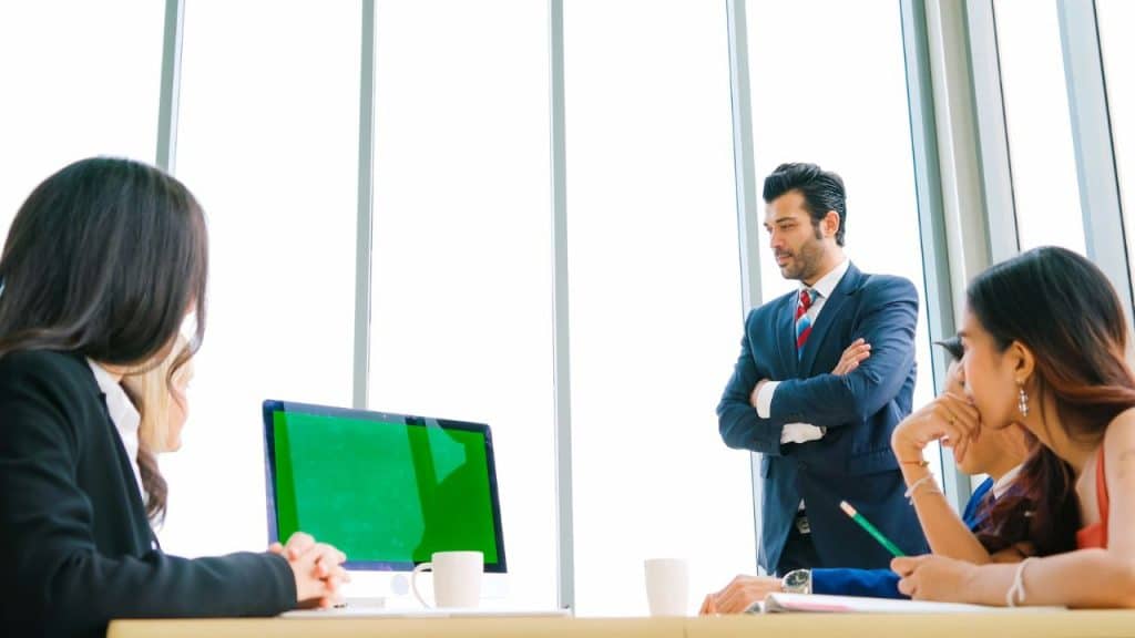 A serious businessman in a suit stands, with his arms crossed, while looking at three seated colleagues.