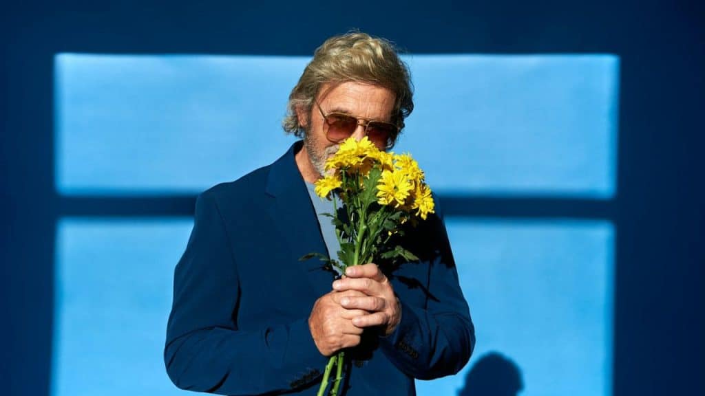 A man in sunglasses and a blue jacket holds and smells yellow flowers.