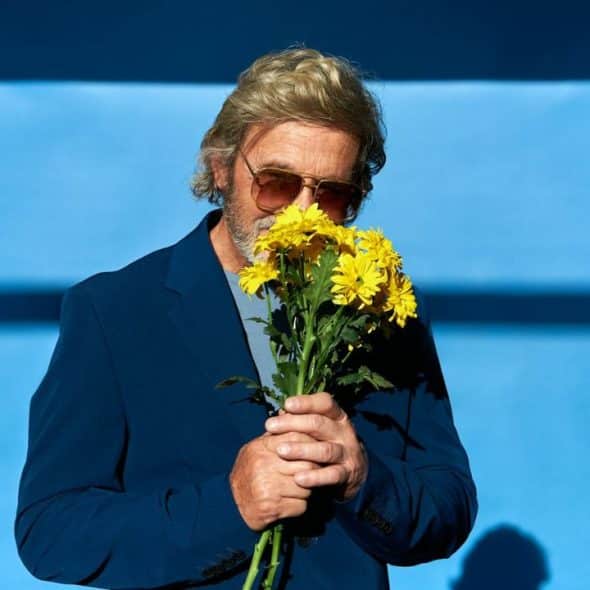 A man in sunglasses and a blue jacket holds and smells yellow flowers.