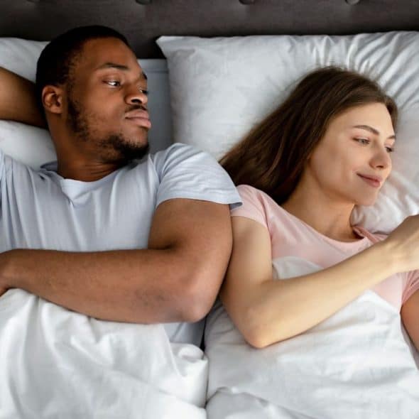 A couple lies in bed. The man looks away from the woman, who is on her phone.