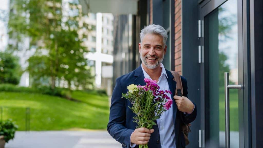 A smiling, gray-haired man in a suit is holding a bouquet of flowers. He is standing outside, next to a building.