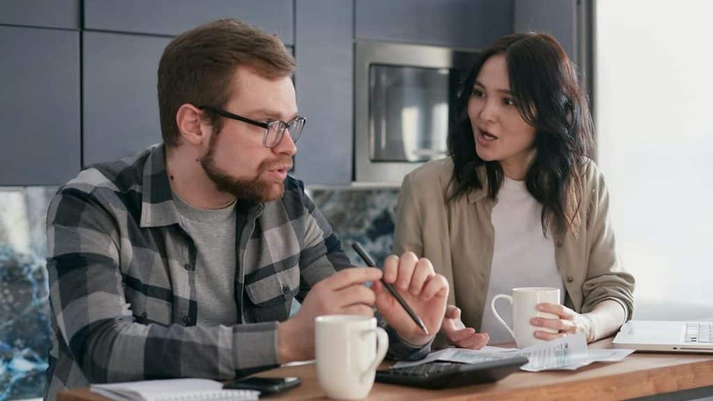 A man and woman sit at a table looking at papers, talking, and drinking from mugs.