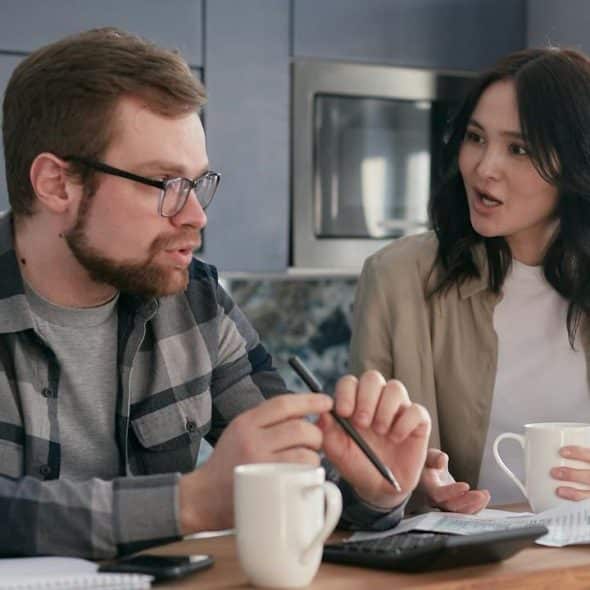 A man and woman sit at a table looking at papers, talking, and drinking from mugs.