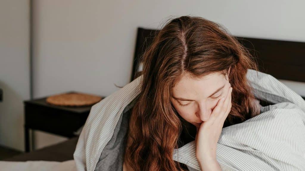 An exhausted woman sitting on a bed with her head in her hand.