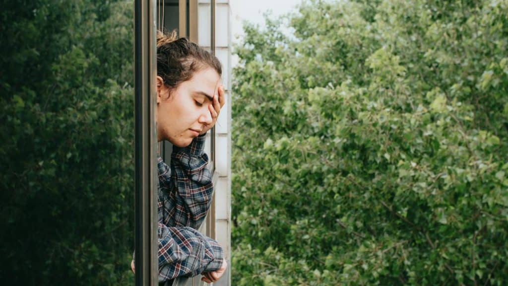 A person leaning out a window with a hand on their forehead.