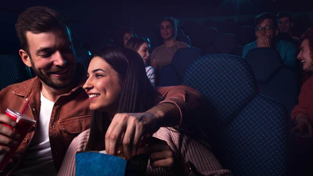 A couple sharing popcorn and drinks while watching a movie in a theater.