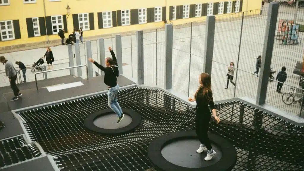 A pair of people jumping on outdoor trampolines surrounded by fencing.
