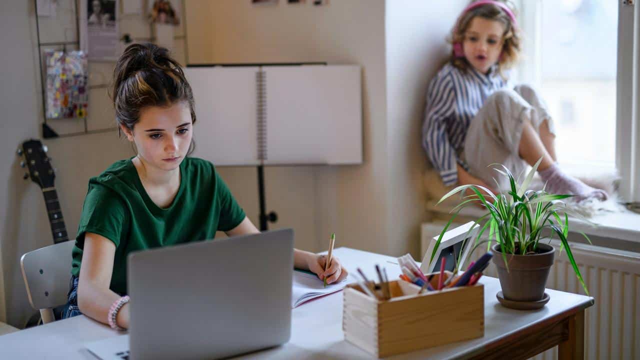 A girl doing homework on a laptop while a child sits on a windowsill.