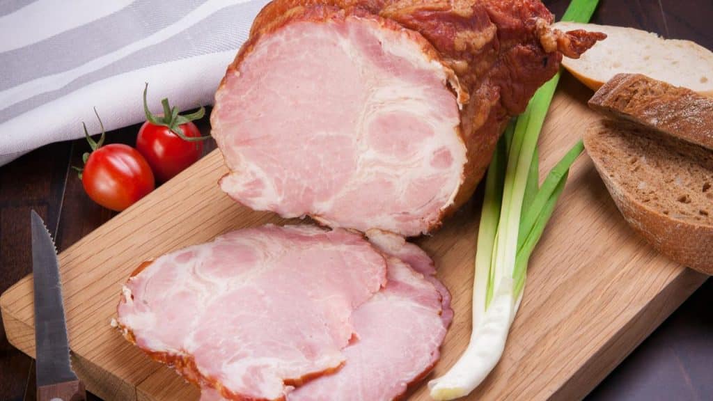 A sliced ham roast on a cutting board with bread and veggies.