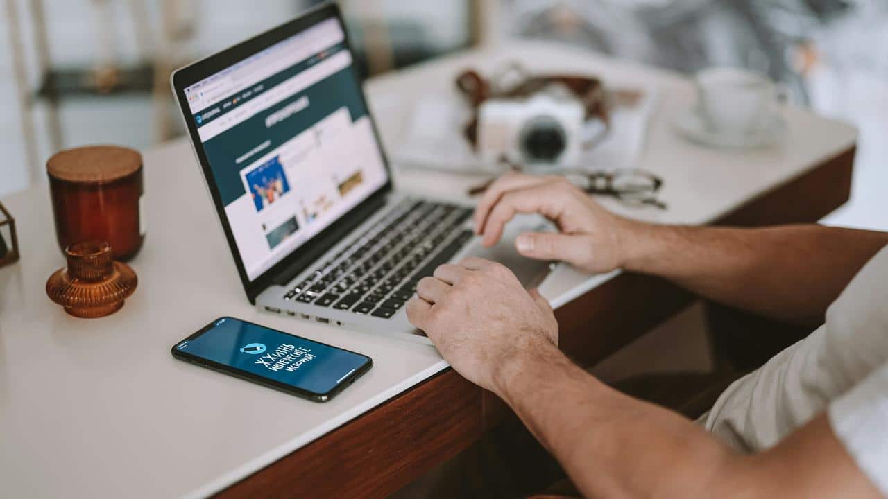 A person's hands typing on a laptop with a phone on the table.