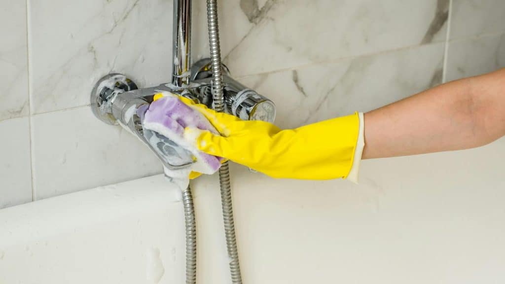 A person cleaning a bathtub faucet with gloves and sponge.