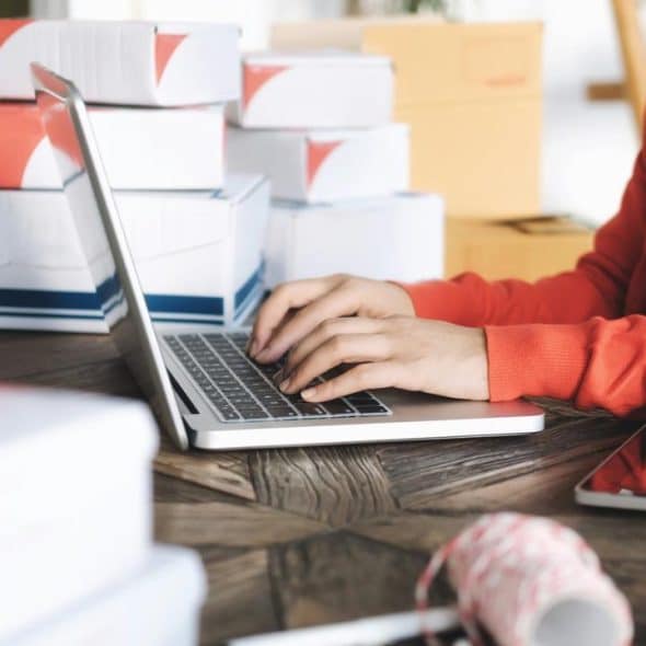 An individual typing on a laptop surrounded by shipping boxes.