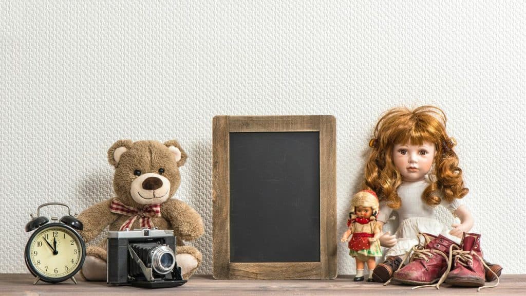 A teddy bear, dolls, shoes, camera, clock, and blank chalkboard on a shelf.