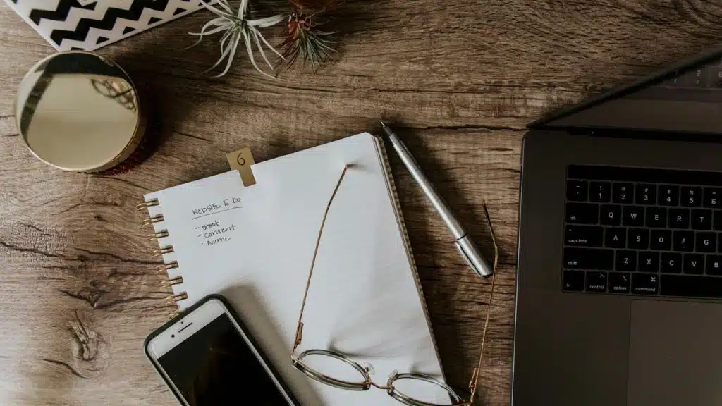 A wooden desk with a notebook, pen, phone, glasses, and laptop.