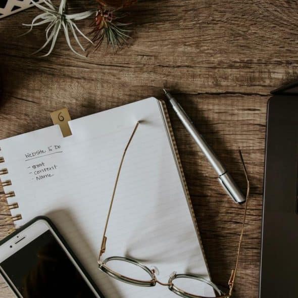 A wooden desk with a notebook, pen, phone, glasses, and laptop.