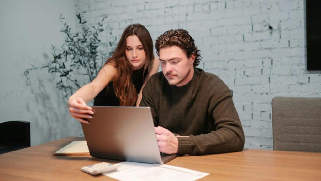 A serious couple works together at a desk, looking at a laptop computer.