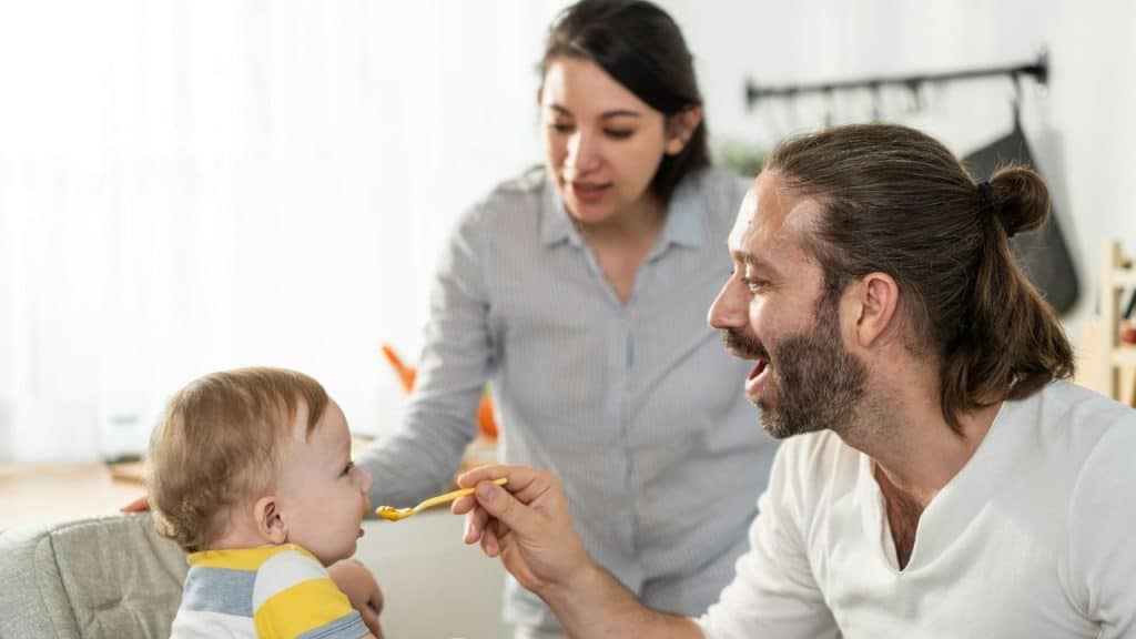 A happy father feeds his baby while his smiling wife watches from behind.
