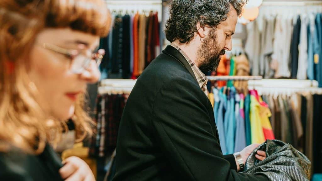 A bearded man looks at clothing in a wardrobe while a woman with glasses stands beside him.