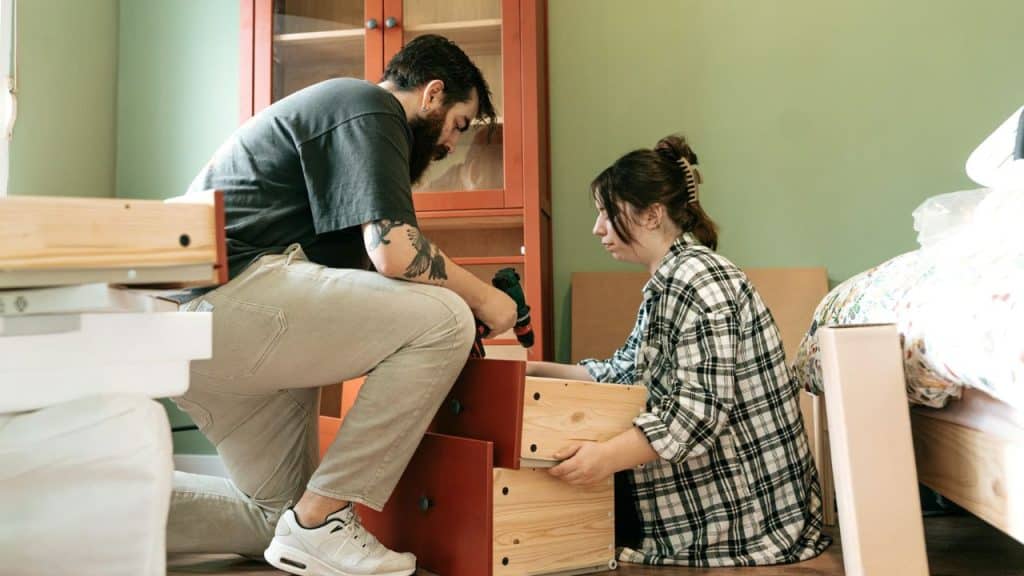 A couple kneels on the floor, working together to assemble a wooden dresser.