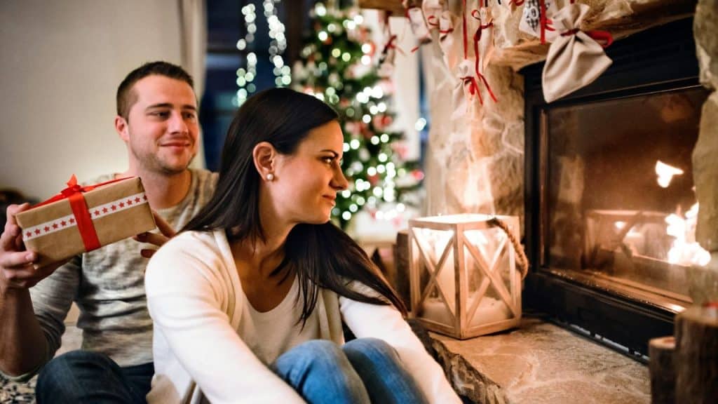 A smiling man holds a gift behind a woman's back while she looks at a fireplace.