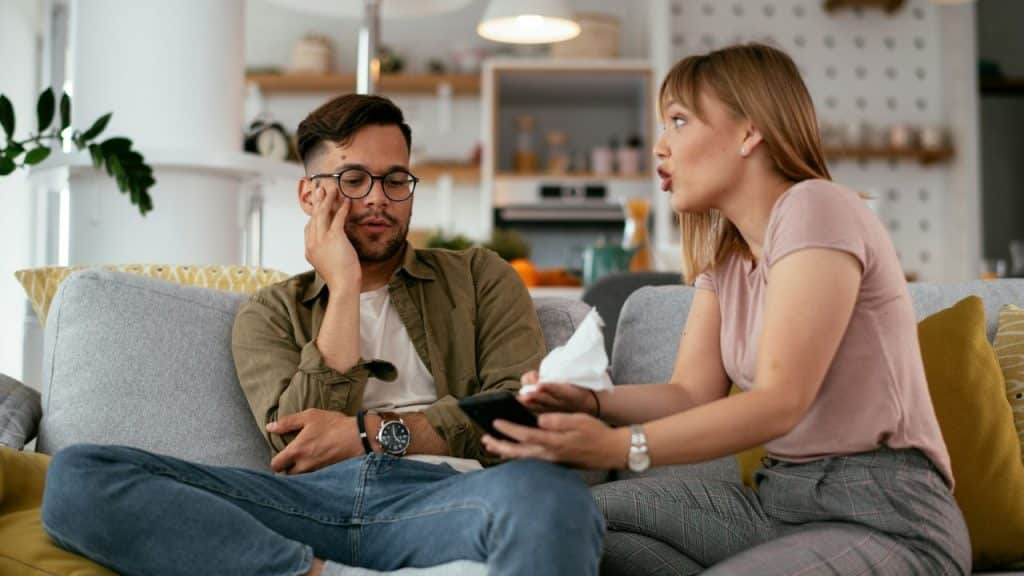 A concerned woman holding a tissue and a phone looks at a tired man sitting beside her.