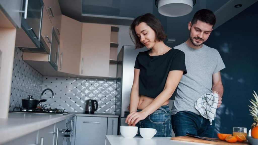 A concerned man with a towel behind his back looks down at a woman at a kitchen counter.
