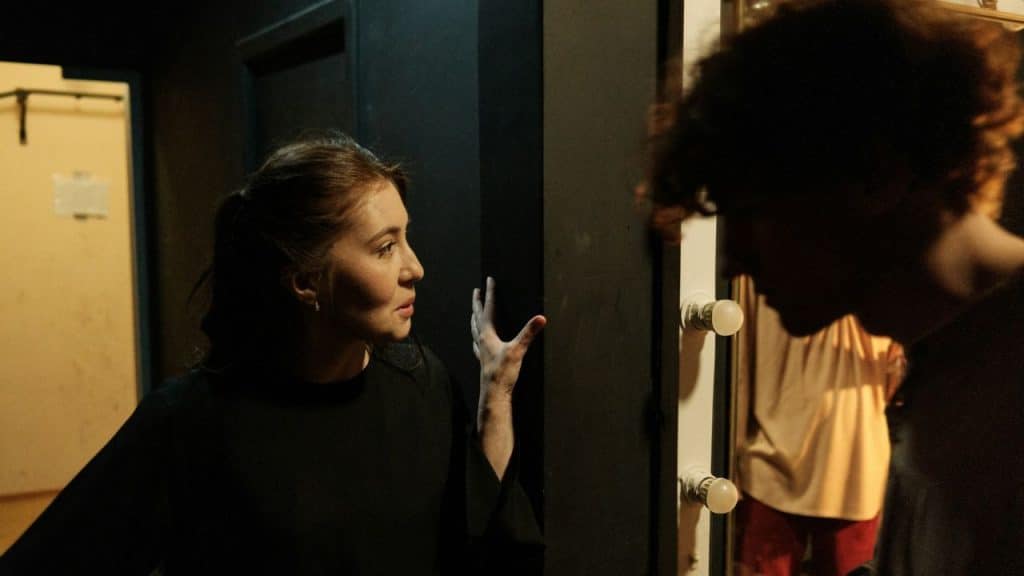A worried woman gestures while speaking to a man in a dark hallway.