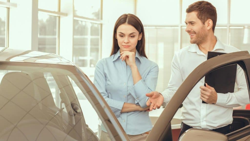 A salesman holding a clipboard talks to a serious-looking woman while showing her a car.