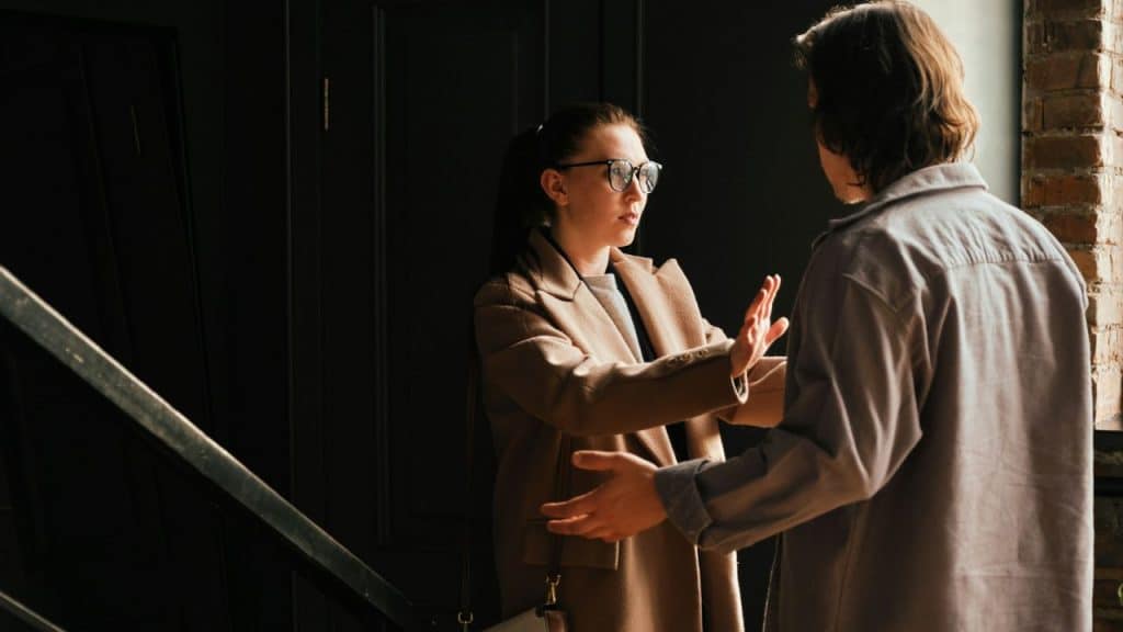 A concerned woman in a beige coat holds her hand up, talking to a man standing across from her.