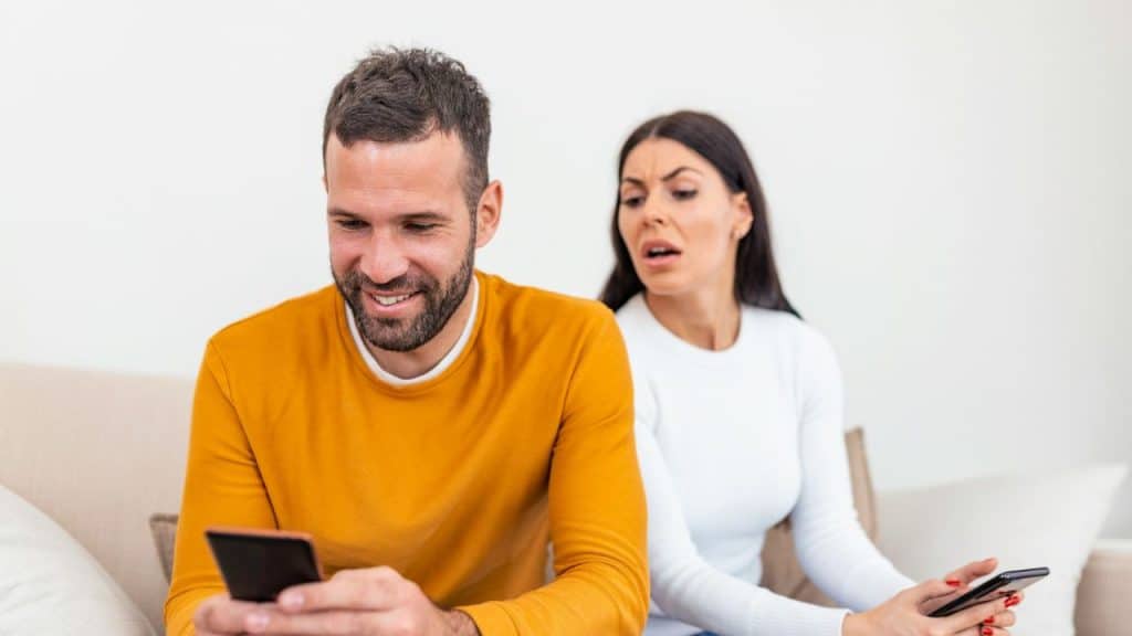 A happy man smiles at his phone while a woman behind him looks on with disapproval.
