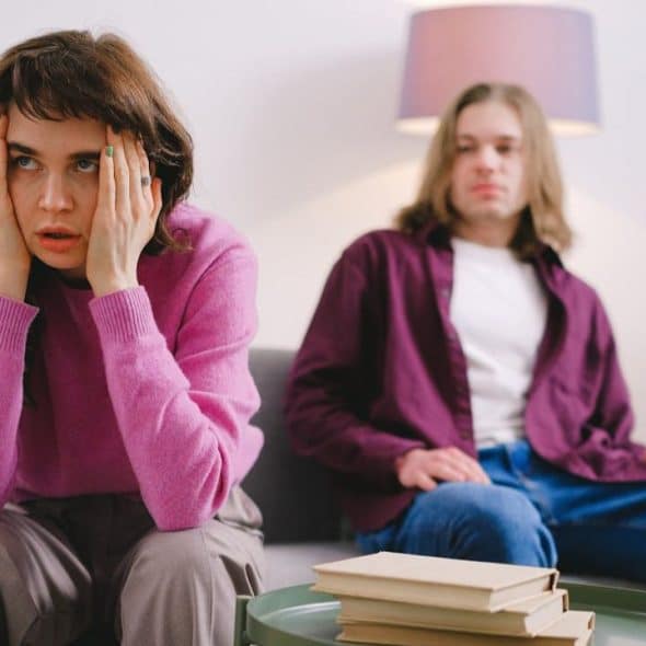 A woman in a pink sweater sits on a couch with her hands on her face.