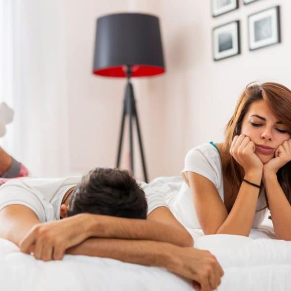 A bored man and woman are lying on a bed with their heads down.