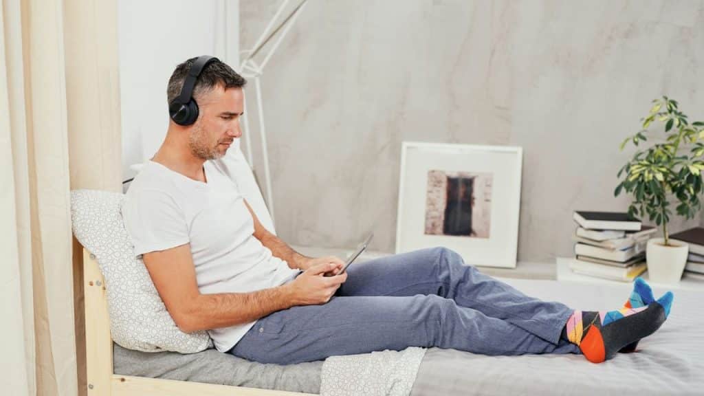 A man wearing headphones is looking at a device while sitting on a bed.