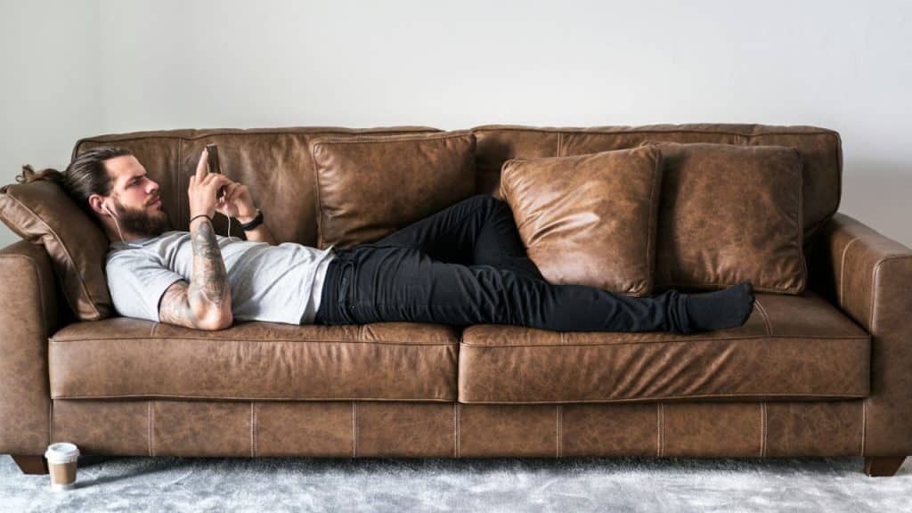 A man with a beard and tattoos is lying on a brown leather couch and looking at his phone.