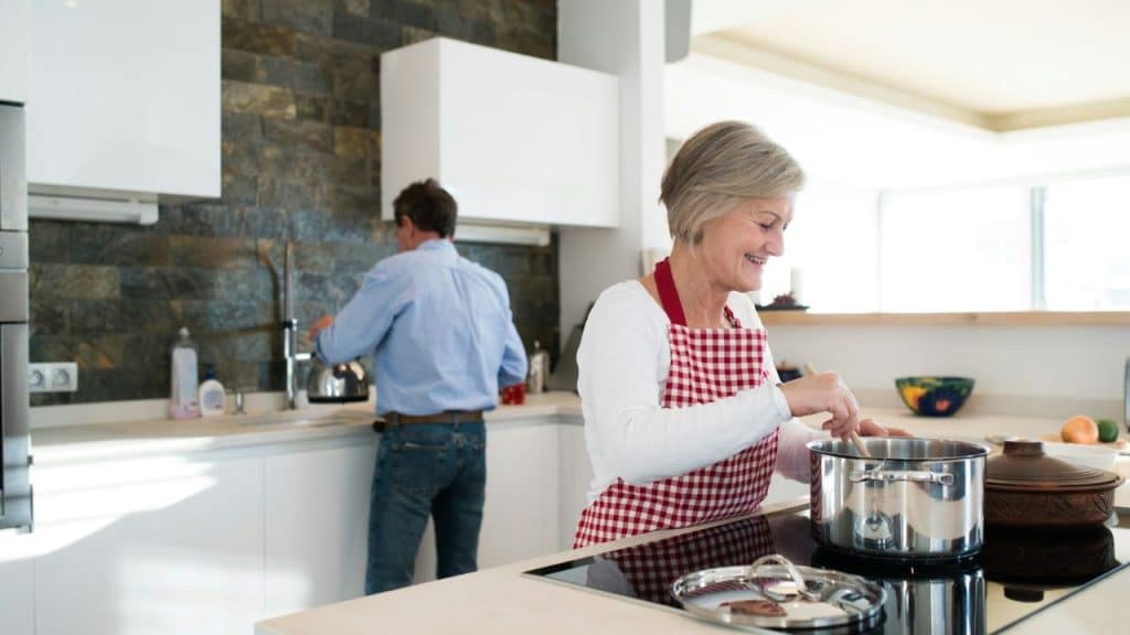 A smiling woman is cooking while a man is washing dishes in the background.