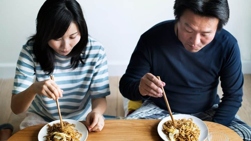 A man and woman are eating noodles with chopsticks from separate plates.