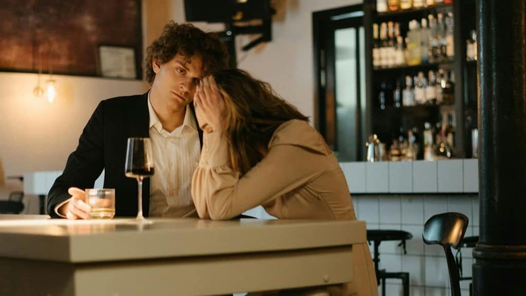 A worried couple sits at a bar, with one holding a glass of wine.