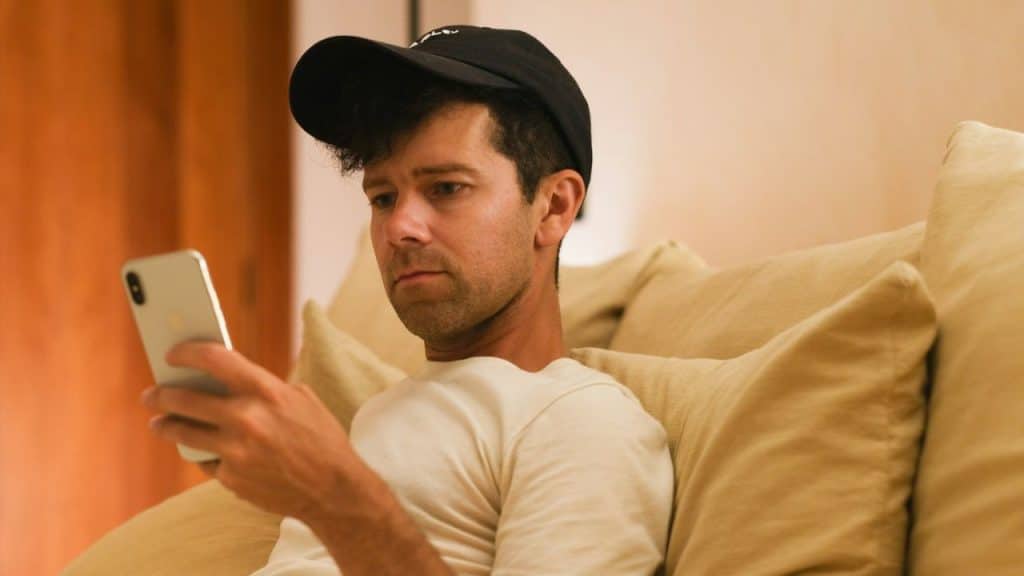 A man with a black baseball cap looks at his phone while sitting on a couch.