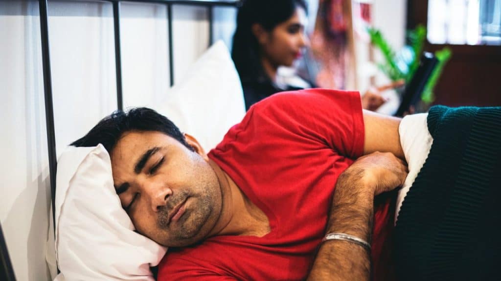 A man is sleeping in the foreground with a woman on a laptop in the background.