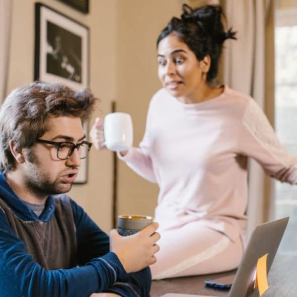 A couple is having a tense conversation, with a man on a laptop and a woman sitting on a table.