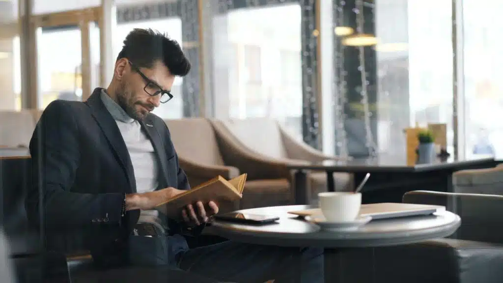 A man in a café reading a book beside a cup of coffee.