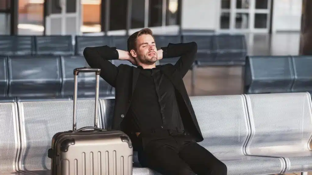 A man relaxing on an airport bench beside his suitcase.