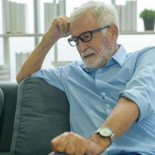 An elderly man with glasses looks troubled while sitting on a couch.