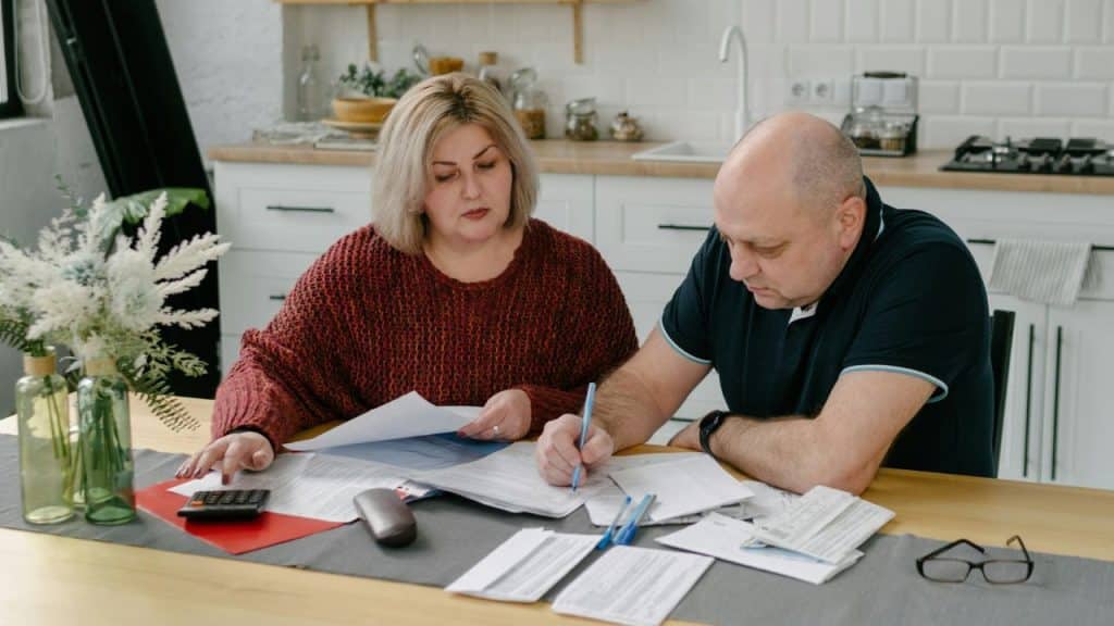 A middle-aged couple at a kitchen table, looking over a pile of bills and documents.