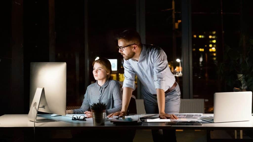 A woman sits at a computer, with a man leaning over her shoulder, both working late in an office.